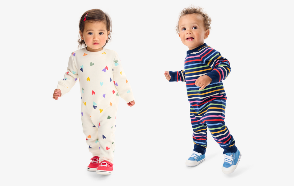 2 babies standing smiling at the camera wearing rainbow cozy rompers.